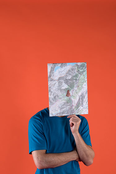 jeune homme dont le visage est caché par une carte IGN seul un trou au centre de la carte laisse sortir son nez.