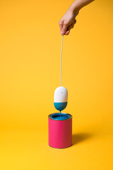 Une main tient une souris d'ordinateur Apple en suspension par le fil, la souris trempe dans un pot de peinture. Design des soeurs siamoises