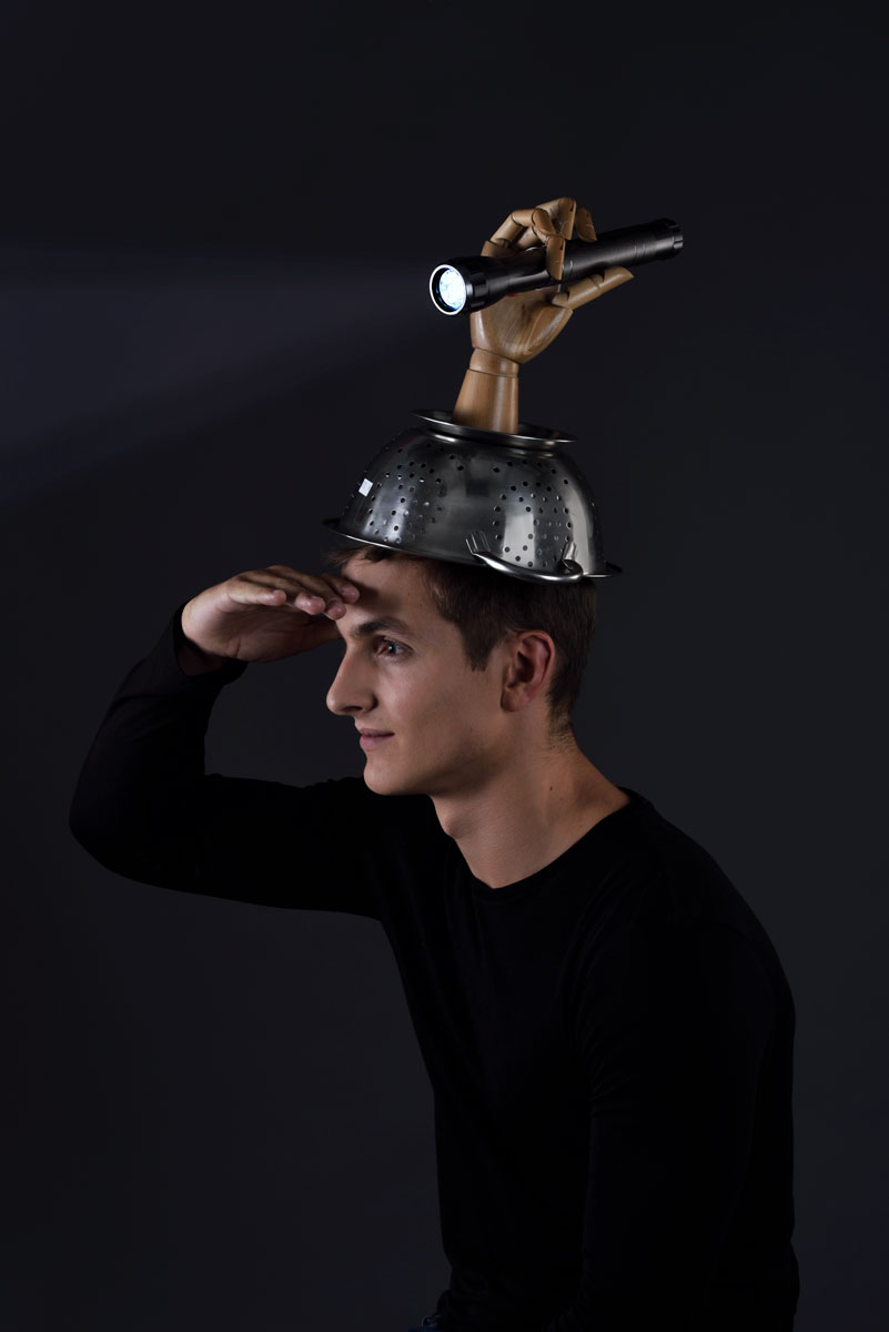 Un jeune homme dans le noir, main au dessus des yeux regarde au loin, il porte pour s'éclairer une passoire sur la tête avec une lampe torche.