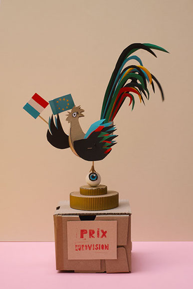 Un trophées avec un texte Prix eurovision, un coq en papier perché sur un oeil sont sur un socle. Design les soeurs siamoises.