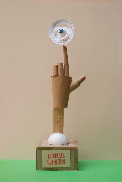 Trophée avec le texte Lentilles surmesure, une main en papier tient un oeil avec mètre de couture autour. Design les soeurs siamoises.